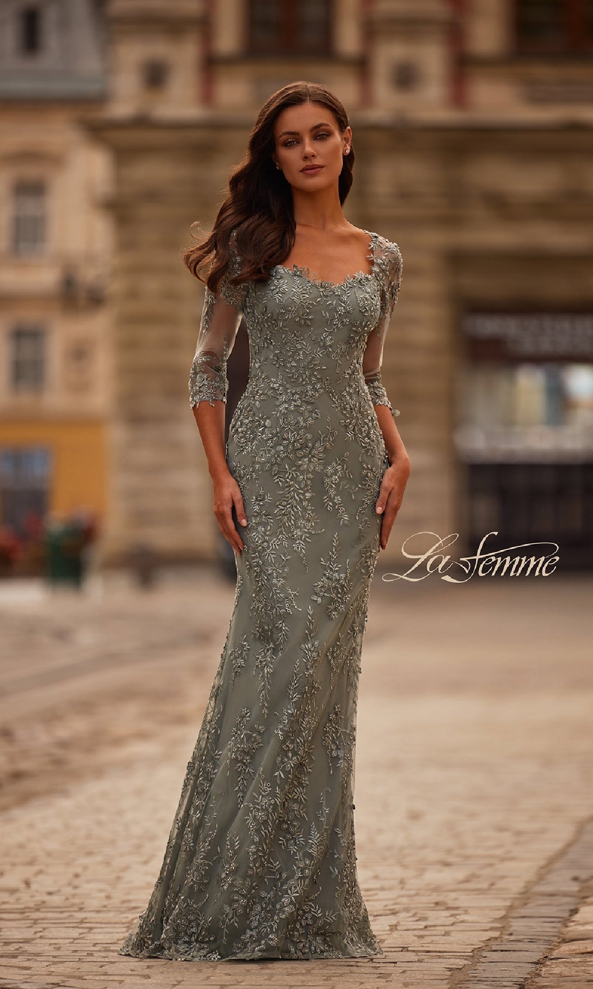 A woman wears the La Femme 31909 long formal dress, featuring a fitted silhouette, lace fabric, three-quarter sleeves, and delicate embroidery as she stands on a cobblestone street.